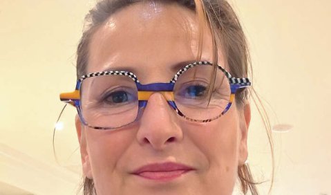 Eine Frau mit dunkelblonden Haaren und einer blau-weiß karierten runden Brille mit gelben Flächen.