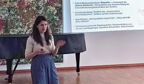 Eine Frau mit hellem Oberteil und lockerer blauer Hose oder Rock hält einen Vortrag in einem Raum mit abgedecktem Flügel.