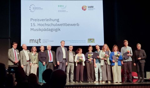 Etwa 20 Personen unterschiedlichen Alters stehen auf einer Bühne mit schwarzem Hintergrund und einer Vortragsleinwand zur Preisverleihung zusammen. Einige halten Blumen und Dokumente.