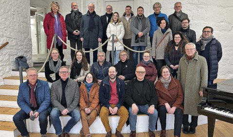 Zwei Dutzend Menschen stehen für ein Gruppenfoto auf einer kleinen Treppe in einem Haus zusammen. Am äußeren Bildrand sieht man einen Flügel.