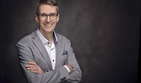 Ein jüngerer Mann in sportlich geschnittenem grauen Anzug mit weißen Hemd.