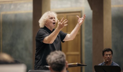 Ein älterer Mann mit weißen Locken hält die Hände energisch gestikulierend vor sich, während er vom Dirigentenpult mit dem Orchester probt.