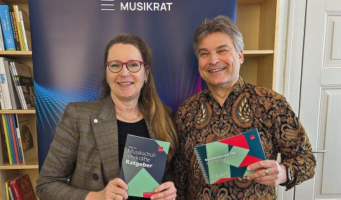 Sie im grauen Blazer, er im gold-braun gemusterten Hemd vor einem Transparent des Deutschen Musikrats.
