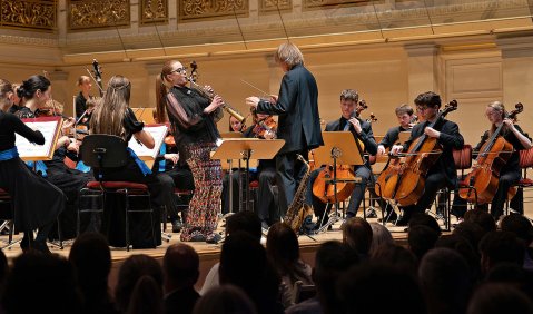 Foto von einem Orchesterkonzert: Die Kameraperspektive ist aus dem Pakett von links auf das Dirigierpult gerichtet und die Solistin gerichtet. Im Hintergrund eine Holzvertäfelung.