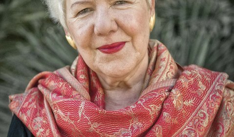 Eine charmant zurückhaltend lächelnde Frau mit kurzen weißen Haaren vor Pflanzen im Hintergrund.