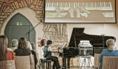 Ein ausgebauter Saal in alten Gemäuern. Darin ein Klavierkonzert mit einem Klavierduo zu vier Händen, deren Hände von oben gefilmt und das Bild mit einem Beamer an die Wand geworfen wird.