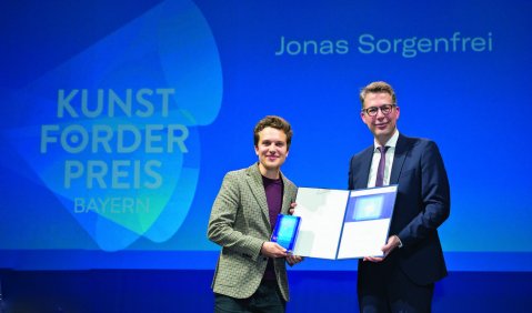 Verleihung des Bayerischen Kuntförderpreis 2023. Der Hintergrund ist Blau und trägt zeigt den Namen des Preisträgers und den Namen des Preises. Davor stehen die in der Bildunterschrift genannten bei der Übergabe des Preises.