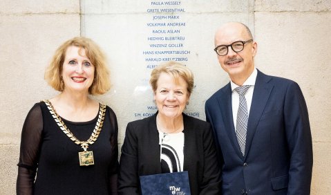 Die drei Menschen mittleren bis höheren mittleren Alters stehen - die Dirigentin in der Mitte - vor einer Namenstafel. Alle festlich gekleidet.