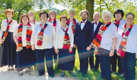 Ein gutes Dutzend Chorsängerinnen mit rot-weiß-schwarzen Schals und ein Chorleiter im Anzug mit Fliege.