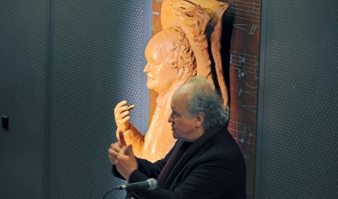 Wolfang Rihm hält eile Rede. Das Foto zeigt ihn schräg von Vorne, hinter ihm an der Wand hängt im gleichen Winkel eine Plastik, die ihn in sehr ähnlicher Pose zeigt.