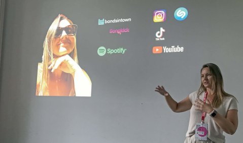 Eine junge blonde Frau mit lockeren weißen Klamotten hält einen Vortrag vor einer weißen Wand, an die diverse Logos von Social-Media- und Musik-Streaming-Anbietern anprojiziert werden.