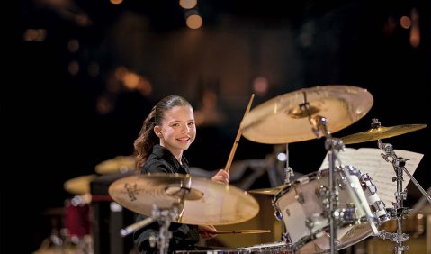 Ein Mädchen mit langen dunkelbraunen Haaren zum Zopf geflochten schaut lächelnd über ihre Drumset hinweg. Der Hintergrund
