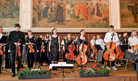 Ein Orchester steht auf einer Bühne. Hinter der Bühne an der Wand prangern sehr große Gemälde. Das Sinfonieorchester trägt teilweise komplett Schwarz, teilweise weiße Oberteile.