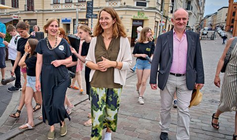 Mehrere Erwachsene Menschen in schick-sommerlicher Kleidung schlendern durch eine Innenstadt. Auch ein paar Jugendliche und Kinder sind zu sehen.