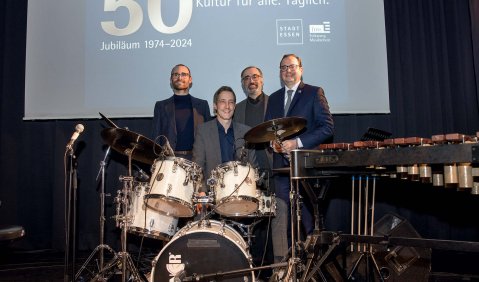 Vier Männer, alle in Anzügen, stehen hinter einem Drumset und neben einem Vibraphon vor einer Leinwand auf der das Jubiläum in goldener Schrift angestrahlt wird.