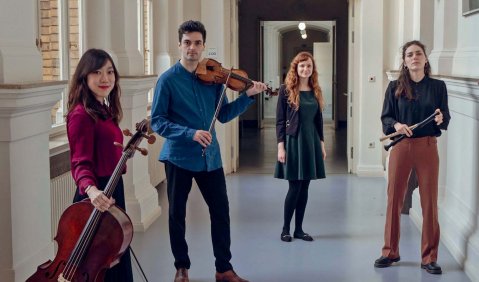 Vier Musiker:innen in unterschiedlichen Kleidung und Pose auf einem Flur.