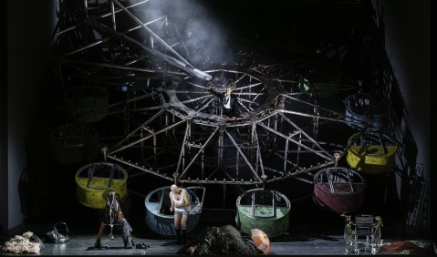 Ein etwa um 120-Grad gekipptes Riesenrad, in den rostigen Gondeln hockt eine Person, eine andere klettert in den Stangen um das Zentrum.