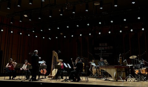 Ein neue Musik-Ensemble mit den erwartungsgemäß gemischten Instrumenten auf einer großen Bühne. Der Hintergrund ist schwarz mit leichten Lichtpunkten der Scheinwerfer.