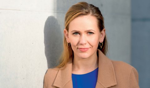 Eine blonde Frau in niedrigem mittleren Alter mit blauem Oberteil unter einem beigen Mantel. Sie steht lächlend an eine weiße Hauswand gelehnt.