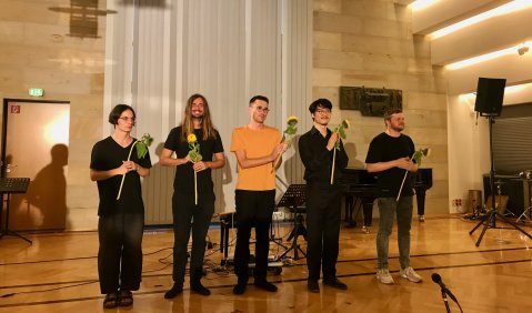 Die 5 jungen Männer stehen mit Sonnenblume für den Schlussapplaus vor dem Publikum im Halberstädter Rathaus. Der Saal ist Modern, Holvertäfelt, im Hintergrund verdecken Lamellen ein Fenster. Die Dachschräge besteht aus Holzstufen mit Leuchtelementen.