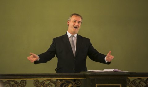 Ein Mann in Anzug und grauen kurzen Haaren steht mit locker ausgebreiteten Armen hinter einer barocken Brüstung. Er singt mit einem Lächeln in den Augen.
