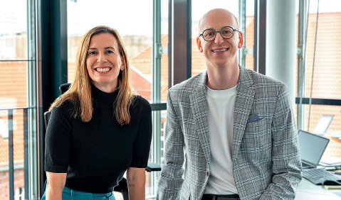 Eine Frau und ein Mann mittleren Alters sitzen mit dynamisch offenem Blick auf einem Tisch in einem komplett verglasten Büro.