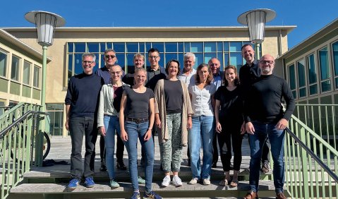 Eine Gruppe von Menschen steht im hellen Sonnenlicht auf einer Treppe vor der Geschäftsstelle.