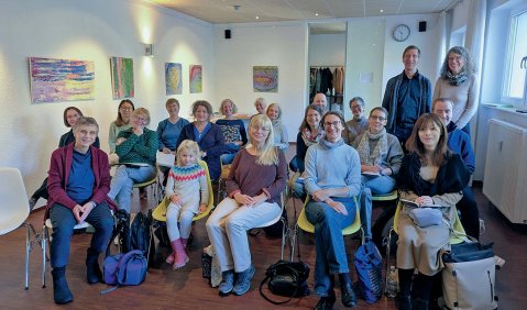 Etwa 20 Personen verschiedener Altersstufen sitzen in einem kleinen Raum wie ein Publikum zusammen.