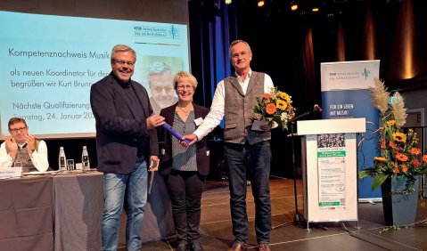 Eine Bühne eingerichtet für ein Podium (ein Redepult, ein Redner:innen-Tisch, dahinter eine Leinwand). Vorne stehen zwei Männer mit einer Frau zwischen sich. Die Männer halten einen Staffelstab, der rechte trägt außerdem einen Strauß Blumen.