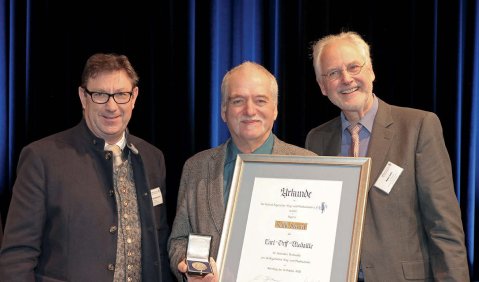 Drei Männer in einer Reihe vor einem blauen Vorhang. Der Linke im mittleren Alter, die beiden anderen etwas älter. Der mittlere zeigt Auszeichnung und Urkunde.