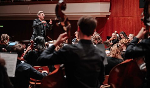 Eine Orchesterkonzert aus der Perspektive hinter den Kontrabässen: Man sieht zwischen den Bässe hindurch Teil des Streichermeeres und vorne (vor den ersten Rängen des Zuschauerraums) einen Dirigenten.
