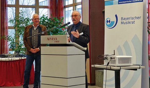 Ein hauptsächlich roter und holzverkleideter Raum mit einem großen Fenster. Rechts im Bild steht ein Beamer und ein Aufsteller des Bayerischen Musikrats, in der Mitte ein Mann in höherem mittleren Alter an einem Rednerpult und links ein Mann ähnlichen Alters etwas neben und hinter ihm..