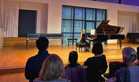 In einem Konzertsaal mit hellen Scheinwerfern und einer indirekt lila beleuchteten Decke spielt ein Mädchen auf der hölzernen Bühne am großen Konzertflügel ein Konzert.
