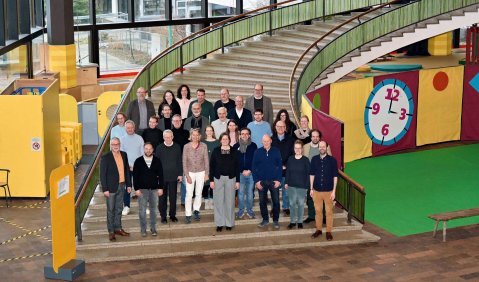 Eine gemischte Gruppe erwachsener Menschen steht auf einer geschwungenen Treppe in einem großen und bunt eingerichteten Foyer.