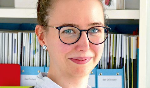 Eine junge Frau mit zurückgebundenen dunkelblonden Haaren steht vor einem Aktenregal und guckt verschmitzt in die Kamera.