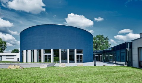 Ein rundes blauies Gebäude mit saftiges Wiese davor und einem Glasfront im Erdgeschoss.