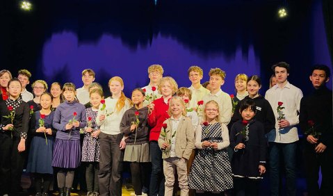 Abschluss-Applaus-Gruppenfoto. Zahlreiche unterschiedlichste Menschen stehen mit einer Blume vor einem Vorhang.