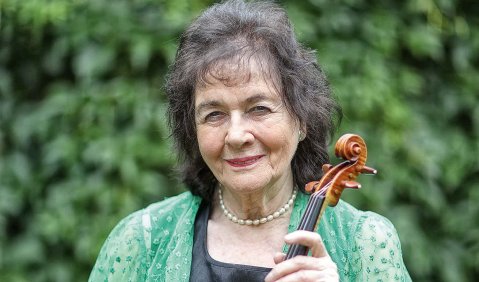 Eine Frau etwas höheren mittleren Alters in grüner Strick/Spitzenjacke mit Violine vor Bäumen.