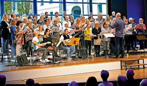 Ein gemischter Chor mit Gitarrenbegleitung während eines Auftritts. Der Chor trägt bunte Kleidung. Im Hintergrund ist eine Glaswand.