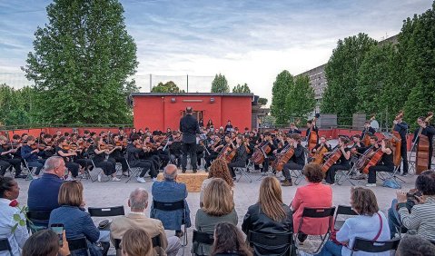 Openairkonzert: Ein Orchester unter einem leicht bewölkten Nachmittagshimmel umzäunt von grünen Bäumen. Erste Reihen des Publikums auf Klappstühlen ist ebenfalls auf dem Foto zu sehen.
