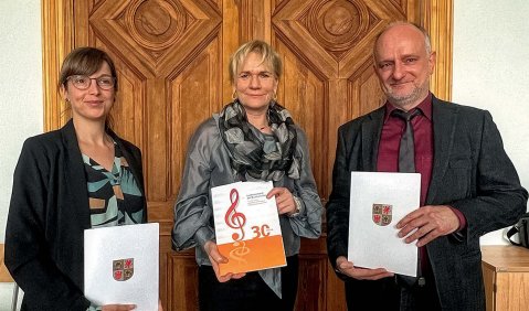 Zwei Frauen und ein Mann stehen jeweils mit Dokumenten in der Hand zusammen vor einer Holzvertäfelung.