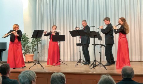 5 Flötist:innen in schwarzen Oberteilen und Hosen bzw. roten Kleidern unter den Oberteilen bei einem Konzert auf einer kleineren Bühne vor einem weißen Vorhang.