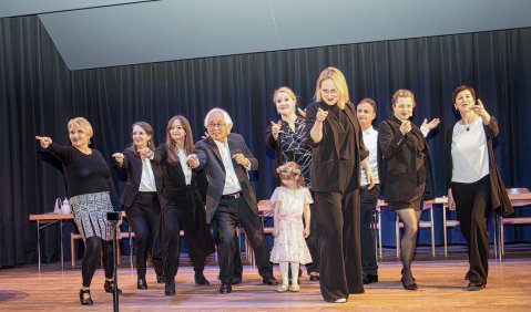 Menschen verschiedener Altersklassen posieren in schwarzweißen Outfits von einer Bühne herunter gegenüber dem Publikum.