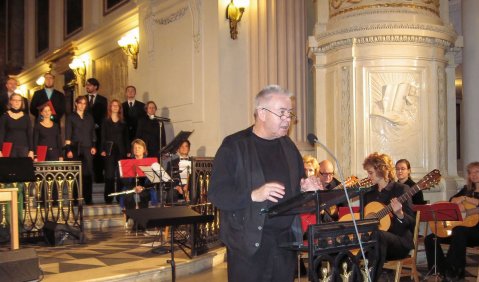 Ein Mann in prunkvolle weiß-schwarz-goldenen Kirche an einem Rednerpult. Im Hintergrund ein Chor und ein kleines Ensemble.