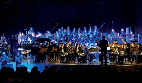 Eine schwer besetzte Bühne mit dem Streichorchester um den Dirigenten in eher gelblichen und die weiter entfernten Musiker:innen in blauem Licht.