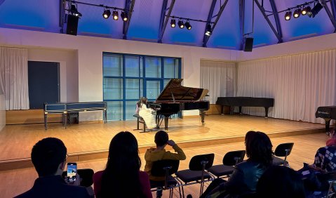 Ein Kammermusiksaal mit niedriger Holzbühne und lila beleuchteten Dachschrägen. Auf der Bühne ein offener Flügel, mit einem Mädchen in weißem Kleid an den Tasten.