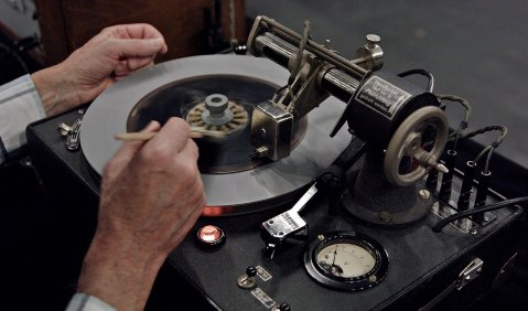 Ein runder Apparat von schräg oben fotografiert. Zwei Hände bedienen das schwarze an einen Plattenspieler erinnernde Gerät. die Recht Hand hält eine Art Stift und bearbeitet einen Metallkranz in der Mitte des Kreises. Über dem Kreis ist ein kleiner Metallarm mit Einstellungsmöglichkeiten und Messangaben.