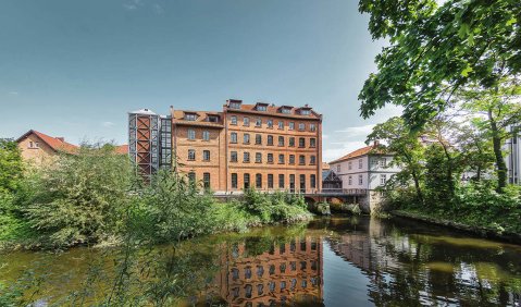 Ein Mehrgeschossiges Backsteingebäude am Wasser mit viel Grün um das Wasser.
