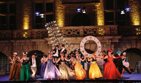 Buntes Treiben vor imposanter Open-Air-Kulisse: Viele junge Operndarsteller:innen in bunten Abendkleidern tanzen vor den alten Gemäuern von Schloss Weikersheim.