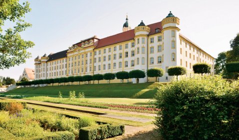 Ein breit angelegtes Schloss mit rotem Dach und wei&szlig;em Putz. Davor ein gr&uuml;ner franz&ouml;sischer Garten.
